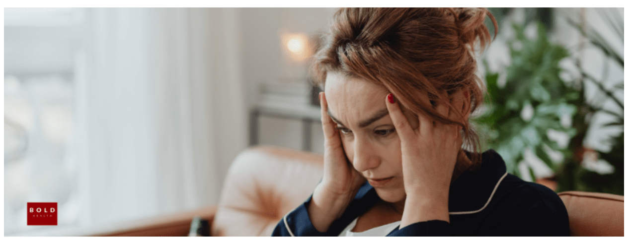 A woman sits on a couch holding her head with both hands, appearing stressed or deep in thought—an image reflecting who can benefit from an IOP in San Diego, with plants and a curtain in the background.