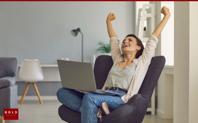 Woman sitting in a chair with a laptop on her lap, stretching her arms upward and smiling in a modern, bright living space.