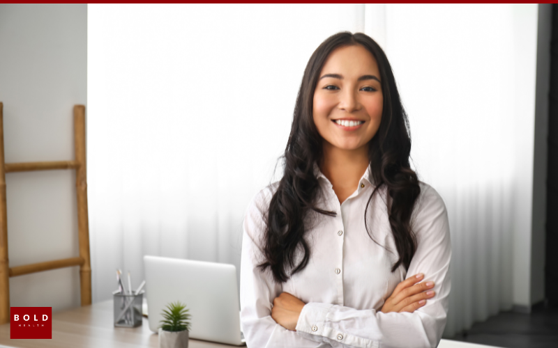 first step to IOP A woman in a white shirt stands with arms crossed, smiling in an office setting with a laptop, plant, and bookshelf in the background.
